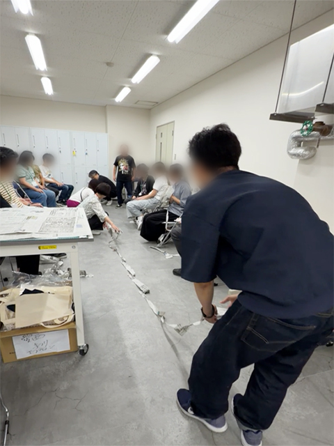 新聞ビリビリゲームの成果として床一面に長く繋がった新聞紙を一直線に並べ、参加者全員で長さを計測している風景。