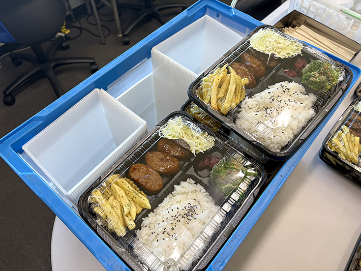青い折りコンの中に完成した白いプラダン製スペーサーを装着し、お弁当パックがすき間なくぴったりと固定されている様子。「見映えが良く使いやすい」とキッチン担当者からも喜ばれた身近な課題解決の工夫。