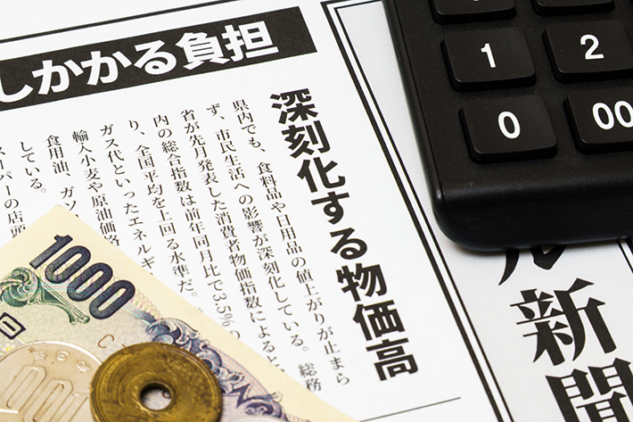 「深刻化する物価高」と書かれた新聞記事の上に、千円札と小銭、黒い電卓が置かれているイメージ写真。世界的な半導体・メモリ高騰や円安の影響によるパソコン価格高騰・導入コストの上昇を解説するビジュアル。