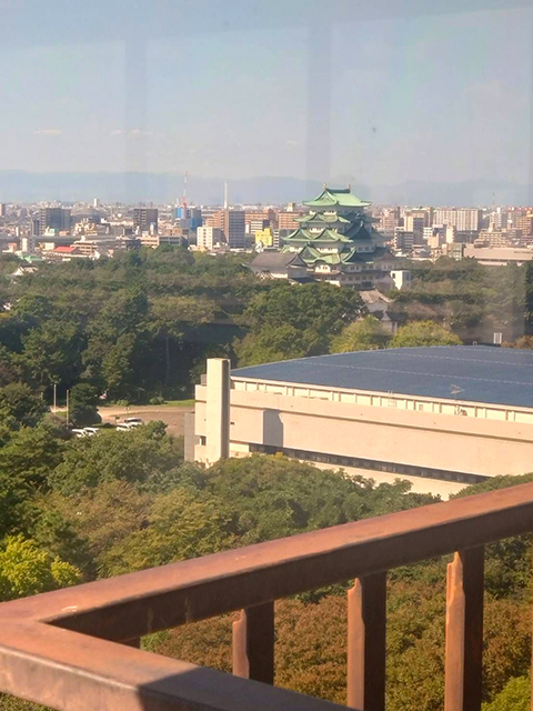 研修会場の窓から眺める、秋晴れの空と名城公園の豊かな緑の中に佇む名古屋城天守閣の風景写真。